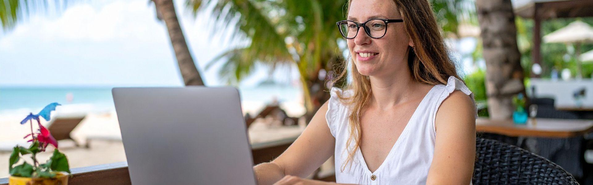 Junge Frau mit Brille und Sommertop sitze am Laptop direkt am Strand unter Palmen