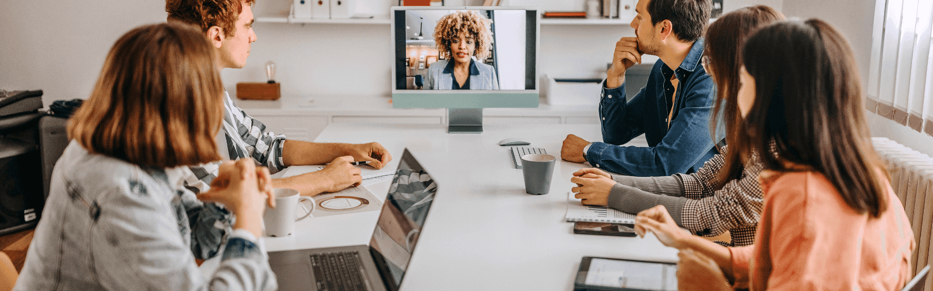 Kollegen sitzen im BÜro und unterhalten sich mit einer Kollegin, die digital dazugeschaltet wurde