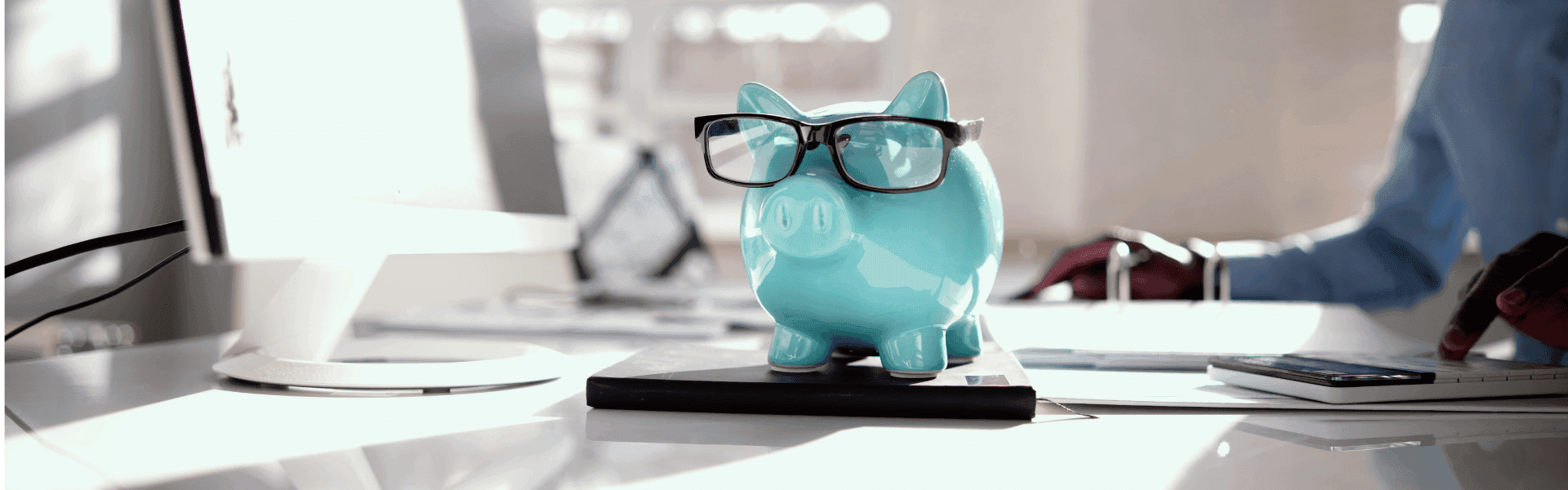 blaues Sparschwein steht auf dem Schreibtisch und hat eine schwarze Brille aufgesetzt
