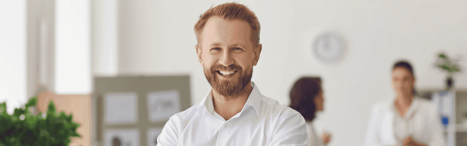 Steuerberater mit Bart lächelt in die Kamera. Im Hintergrund unterhalten sich zwei Kolleginnen. Das Büro ist hell und modern eingerichtet. 