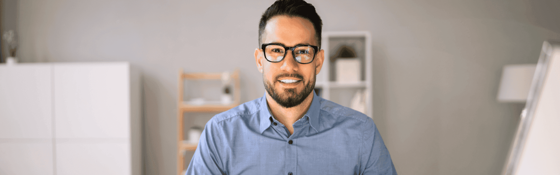 Ein lächelnder Mann mit Brille und Bart trägt ein blaues Hemd. Er sitzt in einem modern eingerichteten Büro mit weißen Regalen und Holzmöbeln im Hintergrund.