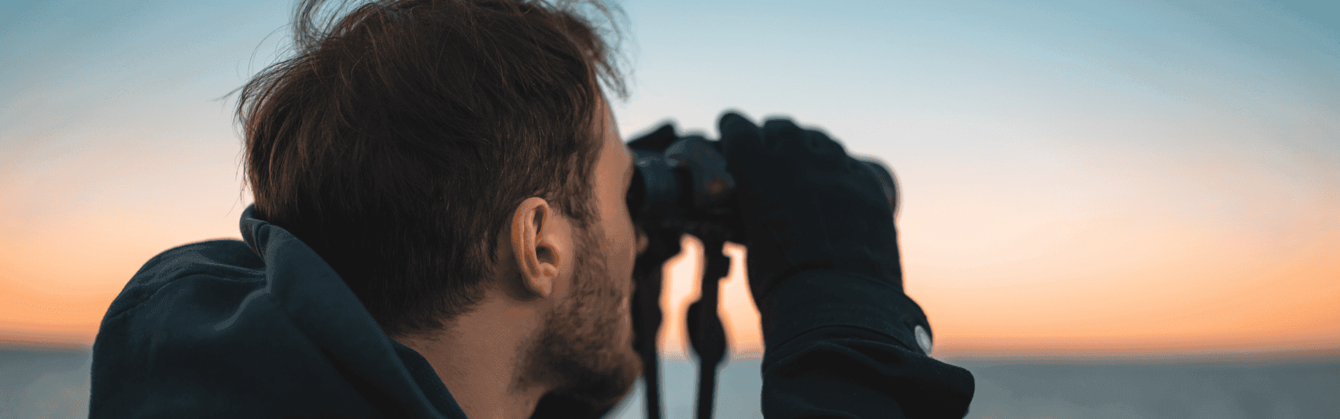 Person schaut mit Fernglas auf das Meer bei Sonnenuntergang.