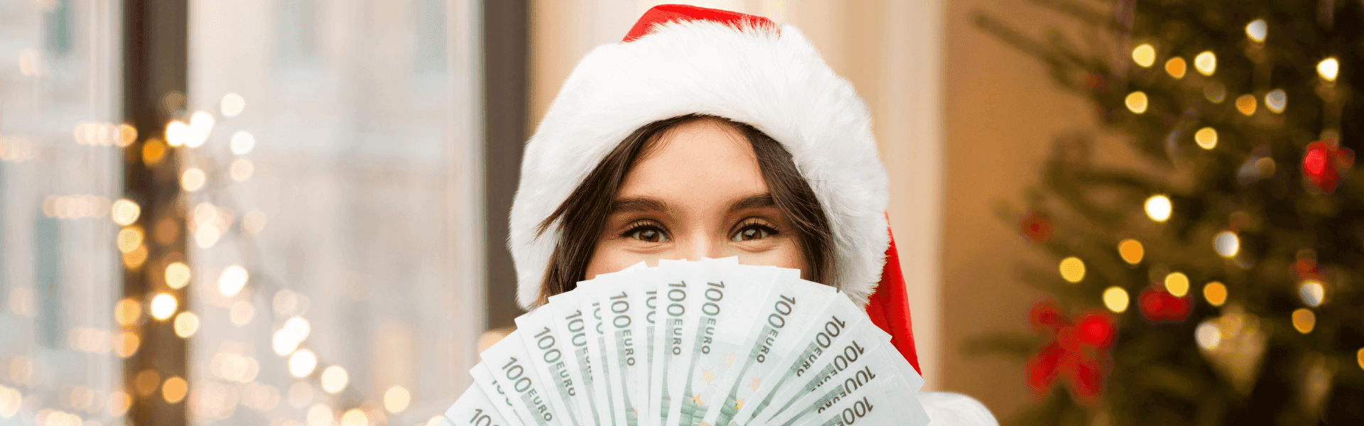 Person mit roter Weihnachtsmütze hält mehrere 100-Euro-Scheine fächerförmig vor sich, im Hintergrund ein festlich geschmückter Weihnachtsbaum und Lichter.