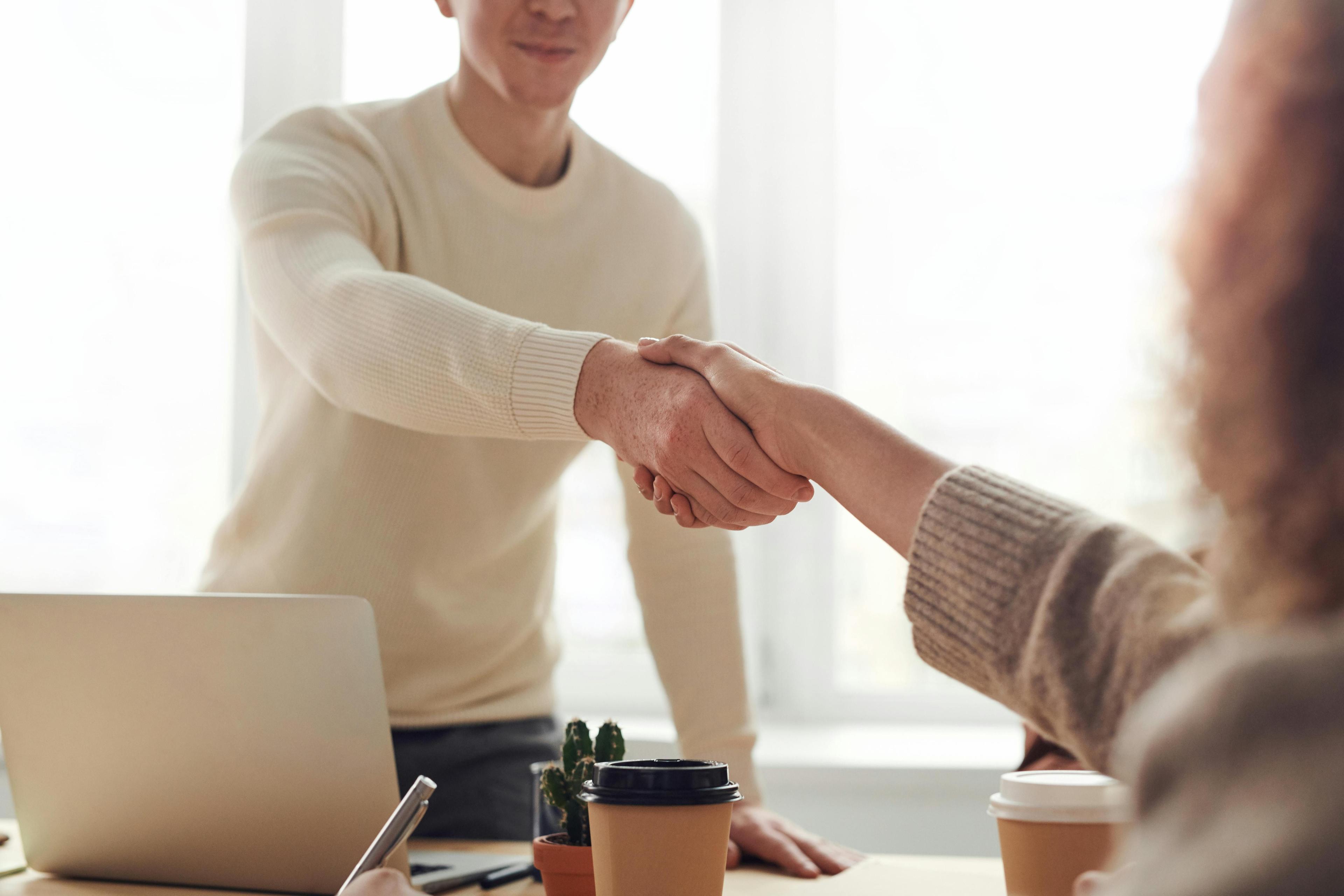 Zwei Personen geben sich in einem Büro die Hand. Auf dem Tisch stehen ein Laptop, Kaffeebecher und ein kleiner Kaktus.