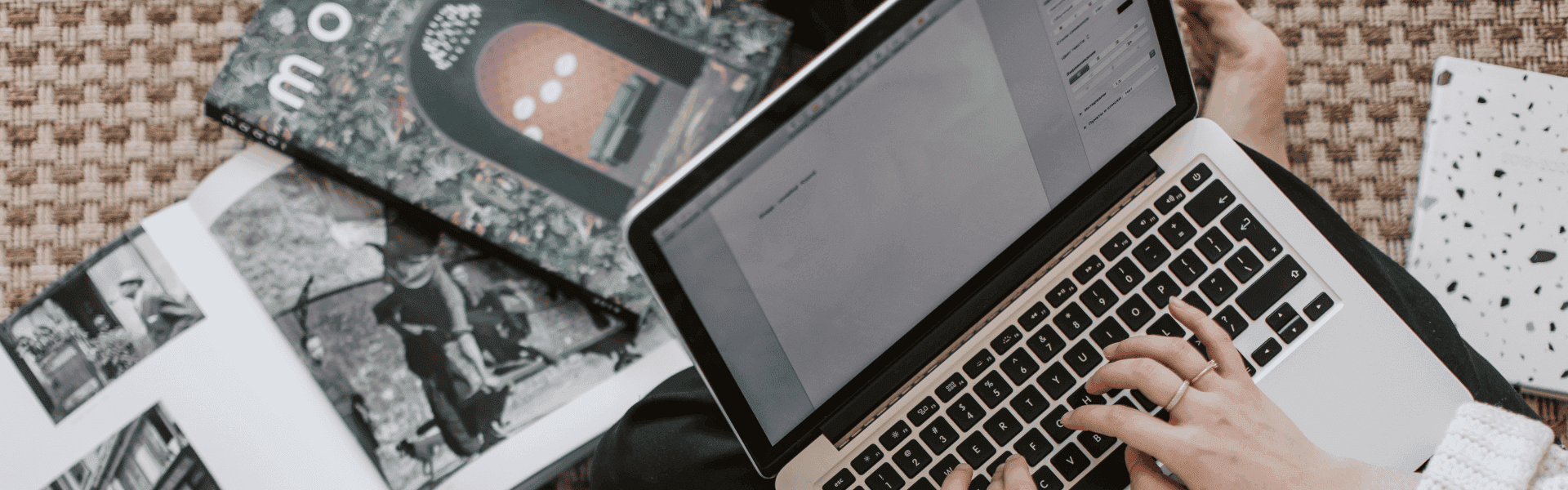 Person tippt auf einem Laptop, umgeben von Zeitschriften und einem Notizbuch auf einer gemusterten Matte.