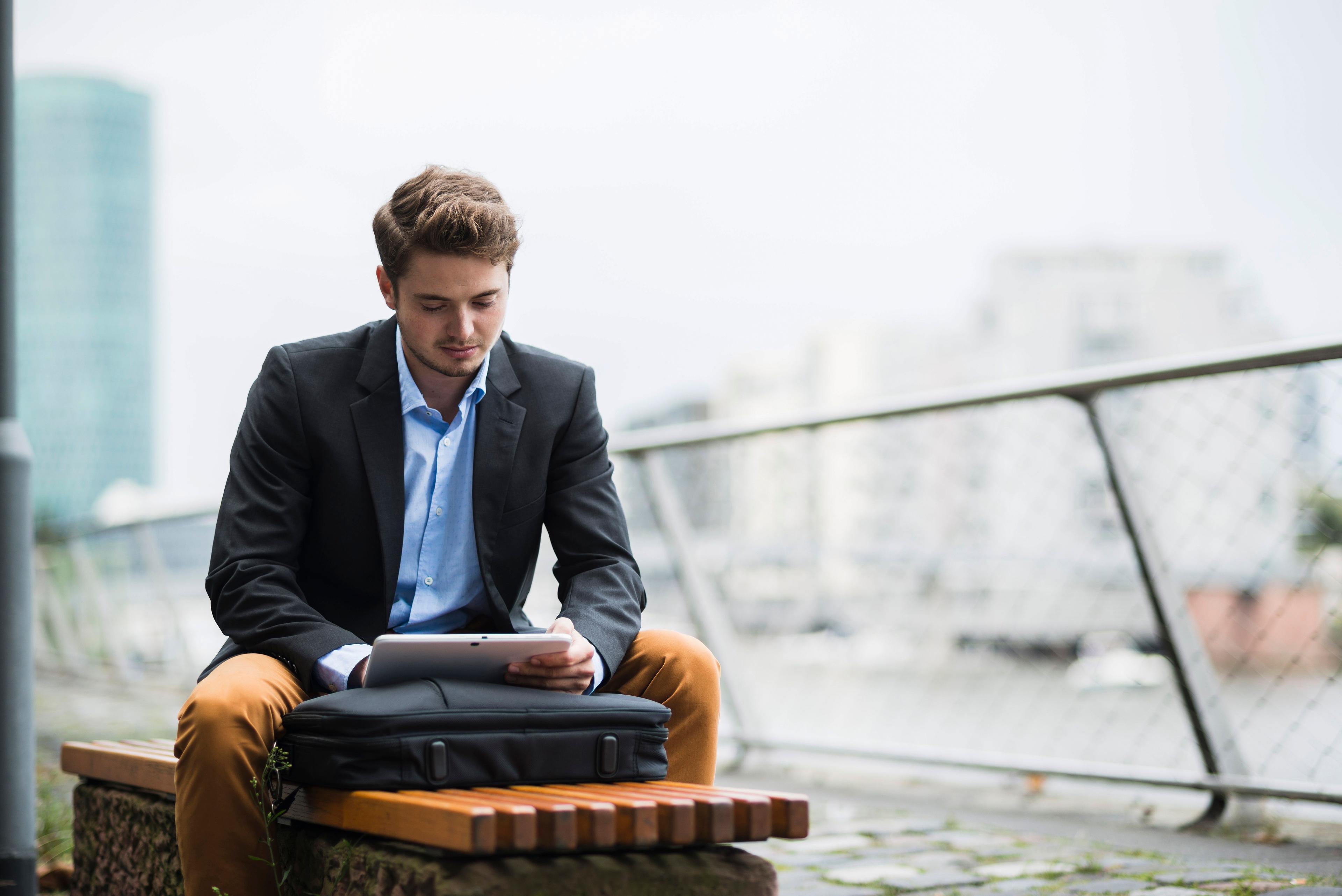 Business Mann sitzt auf einer Bank im Außenbereich eines Bürogebäudes und schaut in seinen Laptop.