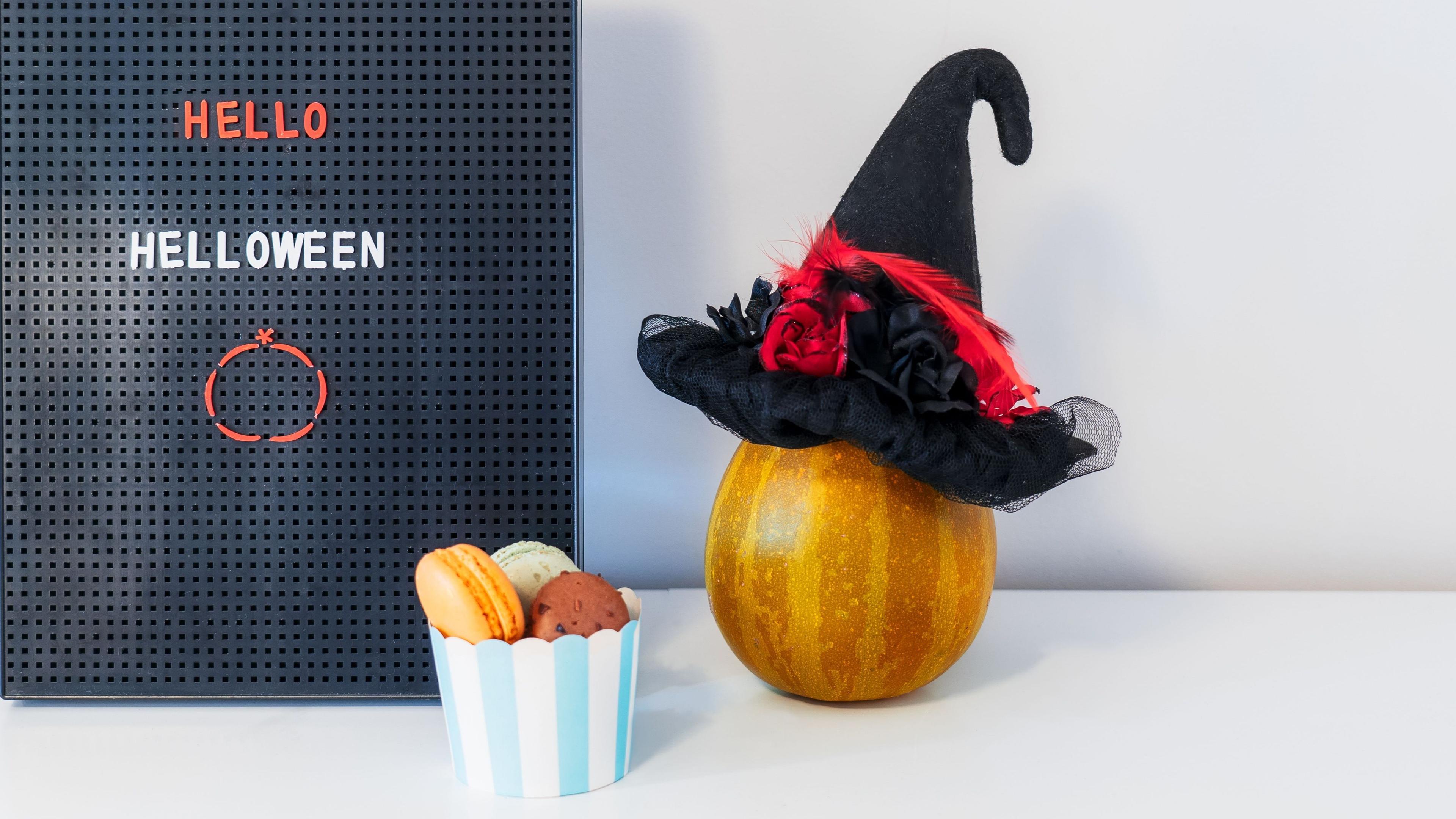 Halloween-Dekoration mit Kürbis, der einen Hexenhut trägt, neben einem Buchstabentafel mit "Hello Helloween". Ein Becher mit Macarons steht daneben.