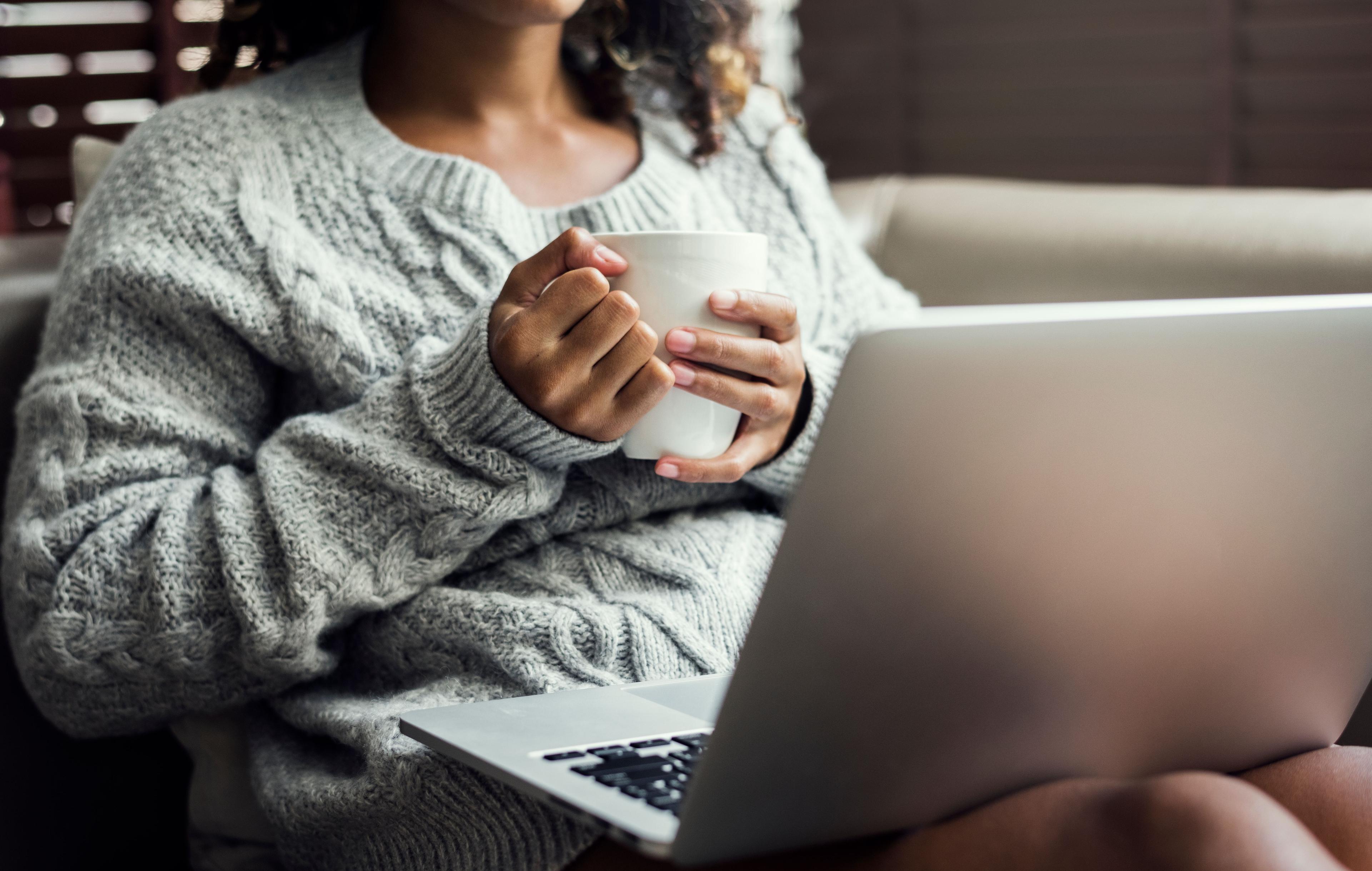 Person sitzt gemütlich auf dem Sofa, hält eine Tasse in der Hand und arbeitet am Laptop, trägt einen grauen Pullover.